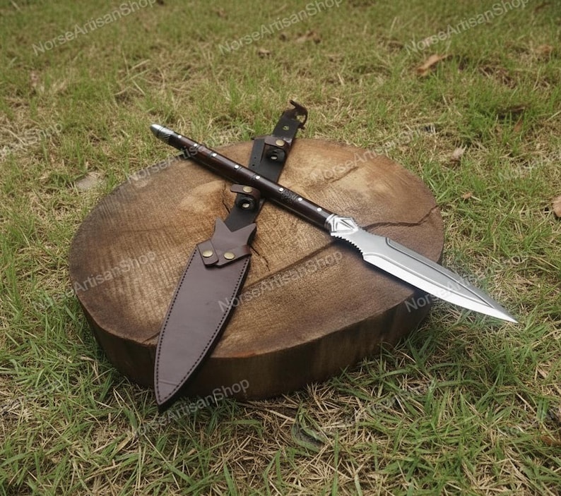 Puede incluir: Una lanza artesanal con una hoja de acero pulido y un mango de madera oscura, apoyada sobre una superficie de madera. Tambi&eacute;n se ven una funda y correas de cuero marr&oacute;n. La lanza est&aacute; dise&ntilde;ada para la recreaci&oacute;n hist&oacute;rica o la exhibici&oacute;n.