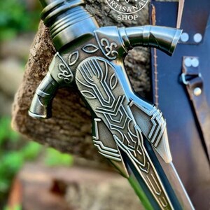 May include: Close-up of a detailed metal sword with a decorative handle. The handle features intricate designs and a silver finish. The blade is partially visible, with a dark, metallic color. The image includes the text "NORSE ARTISAN SHOP".
