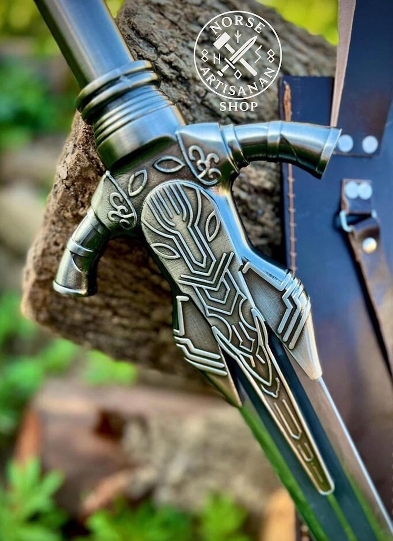 May include: Close-up of a detailed metal sword with a decorative handle. The handle features intricate designs and a silver finish. The blade is partially visible, with a dark, metallic color. The image includes the text "NORSE ARTISAN SHOP".