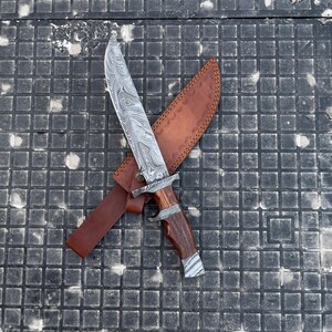 May include: A Damascus steel knife with a wooden handle and a brown leather sheath. The blade has a unique swirling pattern. The handle is a dark brown color with silver accents. The sheath is a lighter brown color.