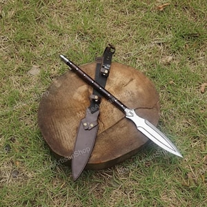 Puede incluir: Una lanza con una hoja plateada y un mango de madera oscura, apoyada sobre un tronco de madera. Tambi&eacute;n hay una funda y un cintur&oacute;n de cuero marr&oacute;n. La lanza est&aacute; dise&ntilde;ada para el combate o la exhibici&oacute;n.