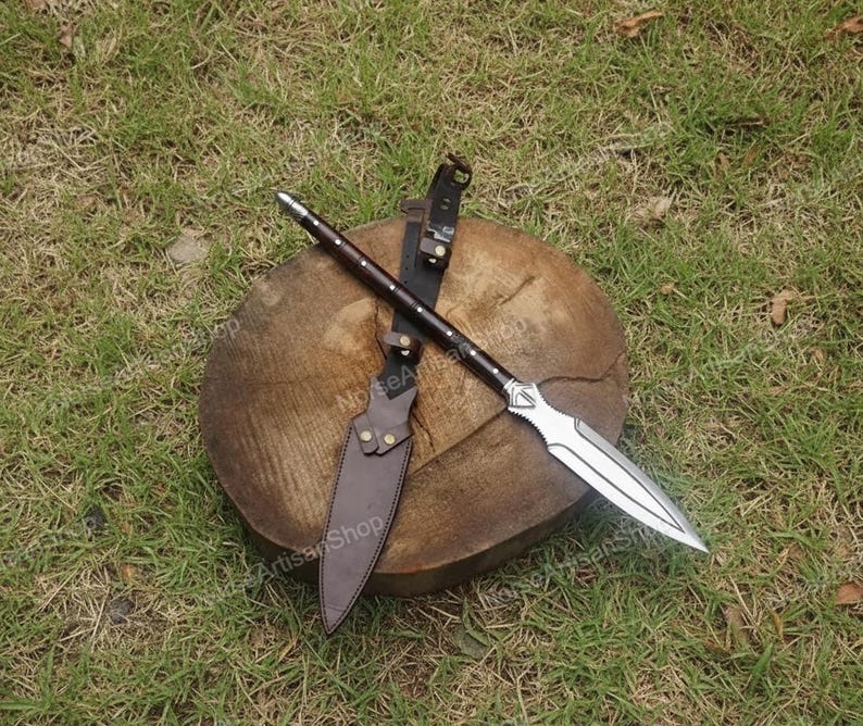 Puede incluir: Una lanza con una hoja plateada y un mango de madera oscura, apoyada sobre un tronco de madera. Tambi&eacute;n hay una funda y un cintur&oacute;n de cuero marr&oacute;n. La lanza est&aacute; dise&ntilde;ada para el combate o la exhibici&oacute;n.