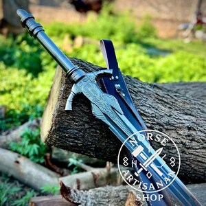 May include: A detailed image of a silver sword with intricate designs, resting on a wooden log. The sword has a decorative hilt and a long, sharp blade. A black leather sheath is visible. The image includes a circular logo with the text "NORSE ARTISAN SHOP".