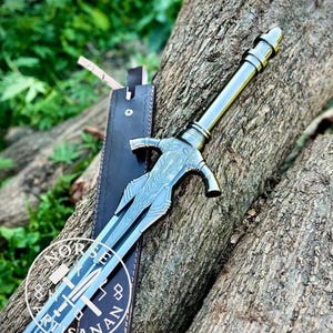 May include: A detailed, silver-colored sword with an ornate handle and blade, resting on a wooden surface. The sword is accompanied by a black leather sheath. The shop's logo is visible on the blade.