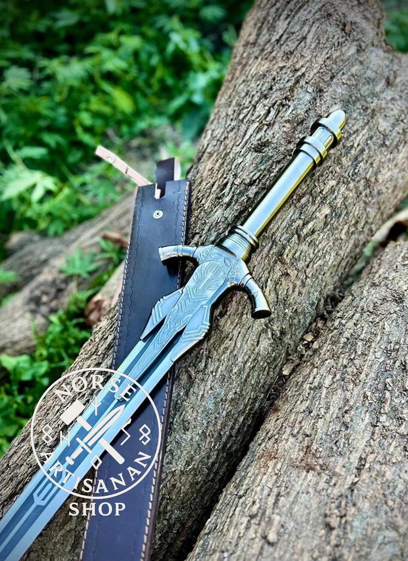 May include: A detailed, silver-colored sword with an ornate handle and blade, resting on a wooden surface. The sword is accompanied by a black leather sheath. The shop's logo is visible on the blade.