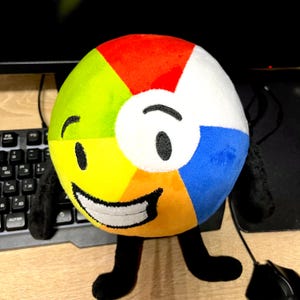 May include: A colourful plush toy in the shape of a beach ball, with segments of red, white, blue, yellow, and green. The toy has a smiling face with black eyes and a mouth, and black arms and legs. It is on a wooden surface.