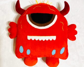 May include: A plush red monster toy with a large eye, horns, and small arms and legs. The monster has a white, scalloped mouth and blue spots. Yellow stitching accents the top of the head.