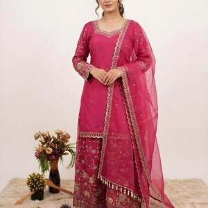 May include: A magenta-colored, embroidered salwar kameez ensemble. The outfit includes a long-sleeved tunic, wide-legged pants, and a sheer dupatta. Gold embroidery adorns the neckline, sleeves, pants, and dupatta edges. The outfit is worn with black and white embroidered flats.