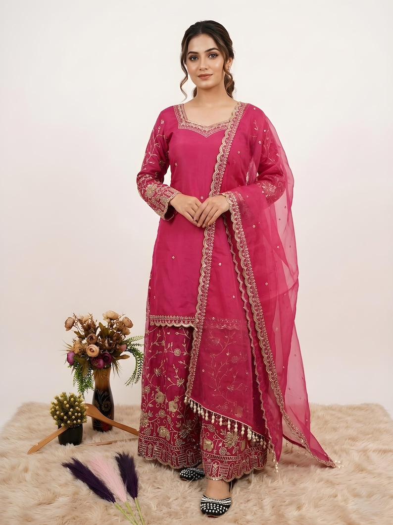 May include: A magenta-colored, embroidered salwar kameez ensemble. The outfit includes a long-sleeved tunic, wide-legged pants, and a sheer dupatta. Gold embroidery adorns the neckline, sleeves, pants, and dupatta edges. The outfit is worn with black and white embroidered flats.