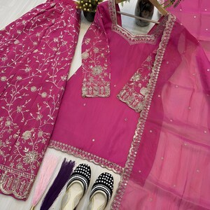 May include: A vibrant fuchsia-colored ensemble featuring a long-sleeved tunic, wide-leg pants, and a sheer scarf. The outfit is adorned with intricate silver embroidery. A pair of black and white polka-dot flats and decorative tassels complete the look. The outfit is displayed on a light-colored surface.