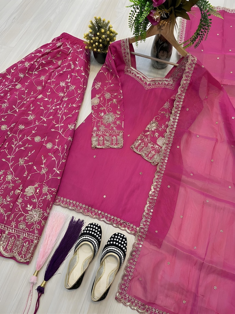 May include: A vibrant fuchsia-colored ensemble featuring a long-sleeved tunic, wide-leg pants, and a sheer scarf. The outfit is adorned with intricate silver embroidery. A pair of black and white polka-dot flats and decorative tassels complete the look. The outfit is displayed on a light-colored surface.