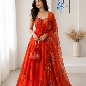 May include: A vibrant orange lehenga set featuring a corset-style bodice with a subtle grid pattern and a floor-length skirt adorned with a dark floral print. A matching sheer dupatta with scalloped edges and sequin floral embroidery is included.