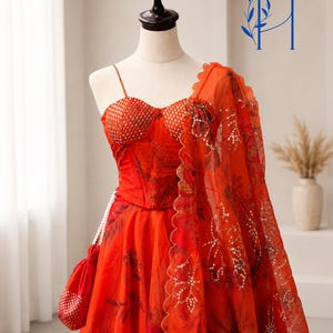 May include: A vibrant orange lehenga set featuring a corset-style bodice with thin straps and a sweetheart neckline, paired with a matching floral print skirt and a sheer dupatta with scalloped edges, accented by a small matching drawstring pouch.