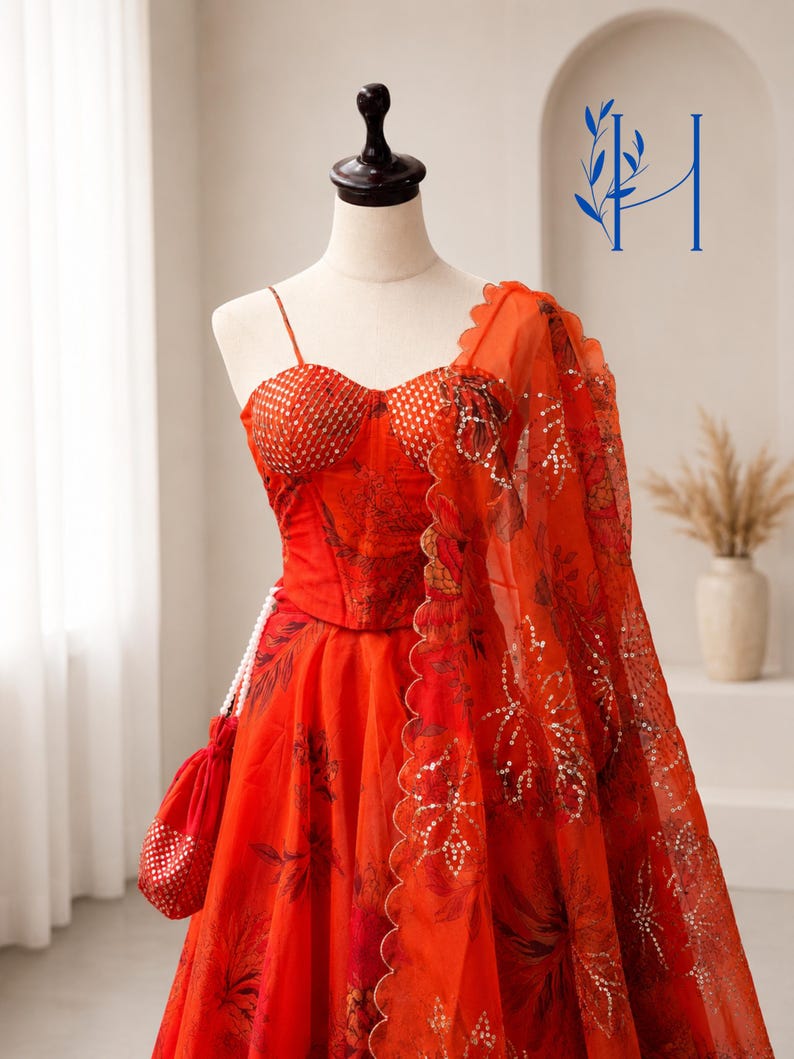 May include: A vibrant orange lehenga set featuring a corset-style bodice with thin straps and a sweetheart neckline, paired with a matching floral print skirt and a sheer dupatta with scalloped edges, accented by a small matching drawstring pouch.