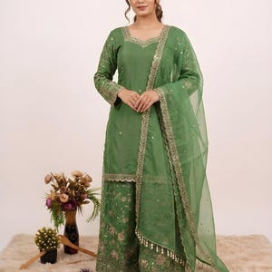 May include: A green embroidered salwar kameez set. The outfit includes a long-sleeved tunic, wide-legged pants, and a sheer dupatta. The ensemble is adorned with gold embroidery and embellishments. The outfit is suitable for festive occasions.