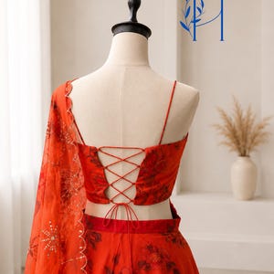 May include: A vibrant red two-piece ensemble featuring a floral print, a sleeveless crop top with a lace-up back closure, and a matching skirt. A sheer red dupatta with scalloped edges and gold sequin detailing is draped over the shoulder.