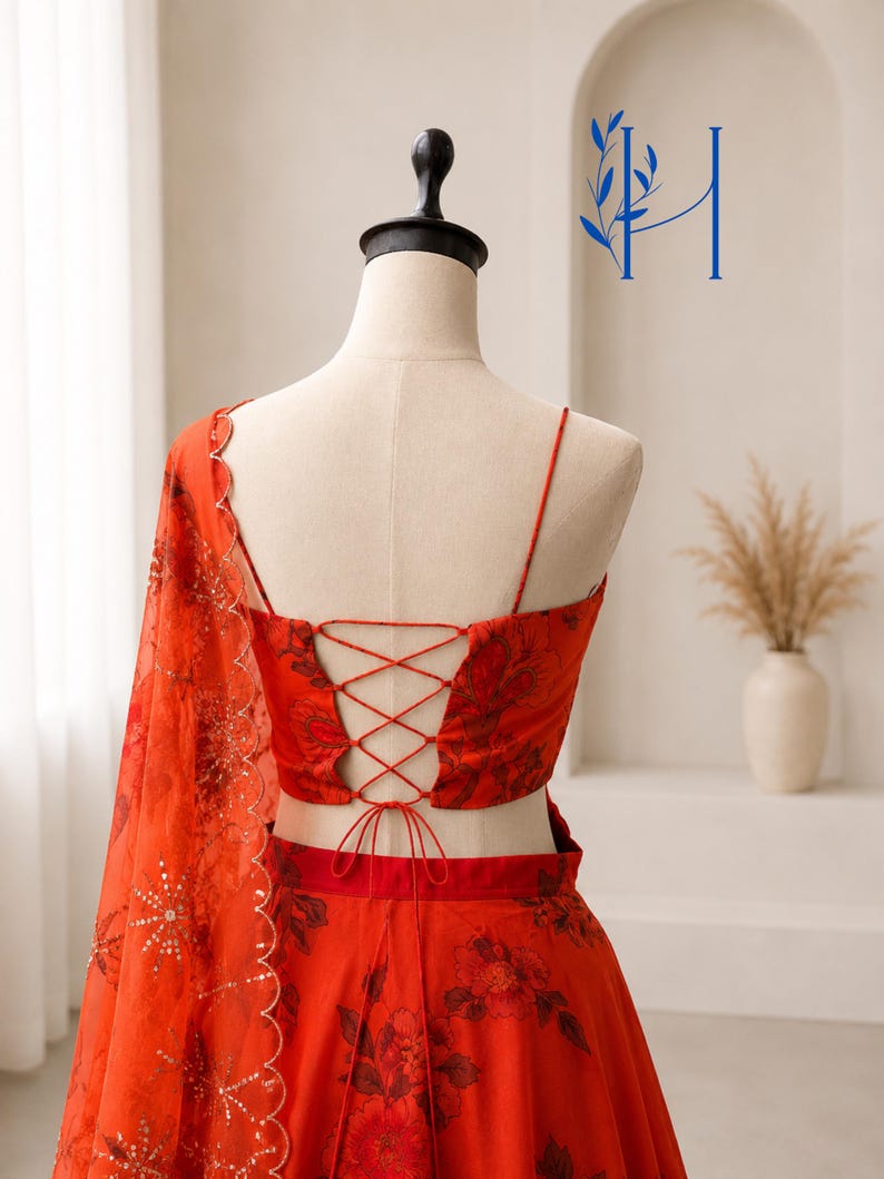 May include: A vibrant red two-piece ensemble featuring a floral print, a sleeveless crop top with a lace-up back closure, and a matching skirt. A sheer red dupatta with scalloped edges and gold sequin detailing is draped over the shoulder.