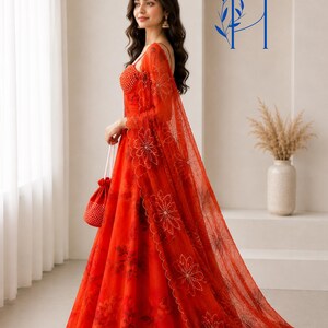 May include: A vibrant red floor-length gown featuring a sheer overlay with intricate floral embroidery and beadwork. The dress includes a matching sheer shawl draped over the shoulder and a small red drawstring pouch bag adorned with white beads.