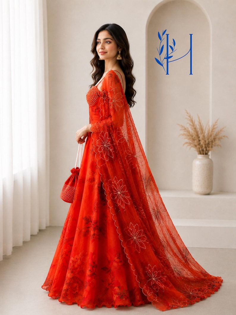 May include: A vibrant red floor-length gown featuring a sheer overlay with intricate floral embroidery and beadwork. The dress includes a matching sheer shawl draped over the shoulder and a small red drawstring pouch bag adorned with white beads.