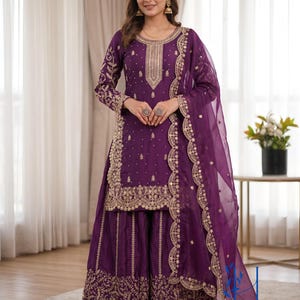 May include: A regal purple salwar kameez ensemble with intricate gold embroidery. The outfit includes a long-sleeved tunic, wide-legged pants, and a sheer dupatta. The embroidery details the neckline, cuffs, and pant hems. The outfit is worn with sandals.