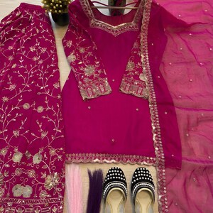 May include: A vibrant fuchsia-colored salwar kameez set with gold embroidery. The set includes a tunic, pants, and a sheer scarf. Black and gold embellished flats and decorative hair strands complete the ensemble. The outfit is displayed on a wooden hanger.