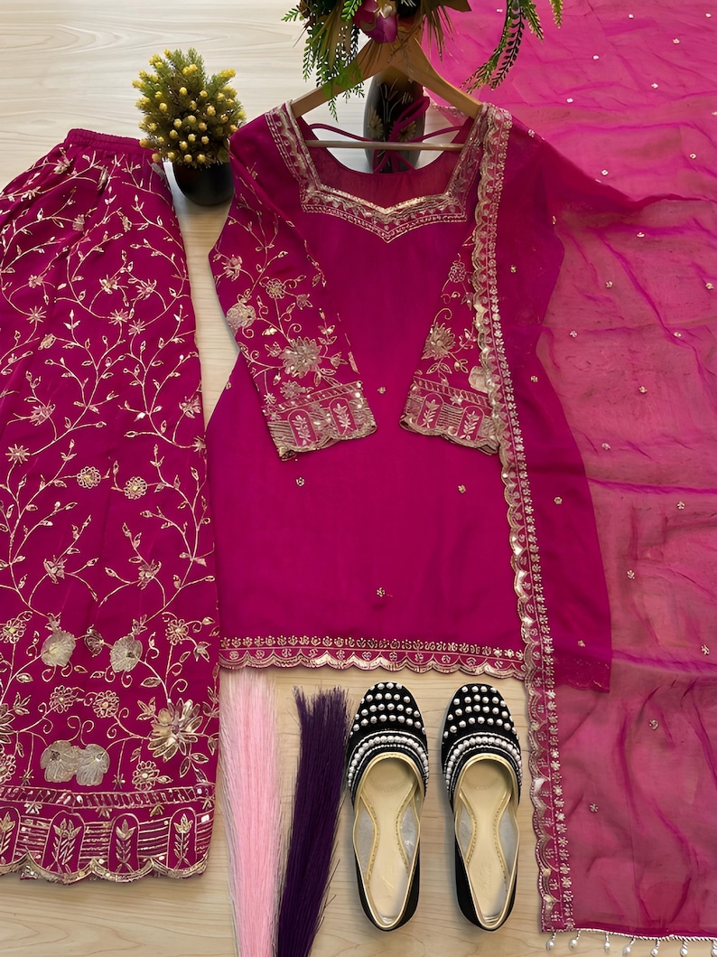 May include: A vibrant fuchsia-colored salwar kameez set with gold embroidery. The set includes a tunic, pants, and a sheer scarf. Black and gold embellished flats and decorative hair strands complete the ensemble. The outfit is displayed on a wooden hanger.