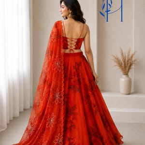 May include: A vibrant red lehenga set featuring a sleeveless crop top with a lace-up back and a full-length skirt with a dark floral print. A matching sheer red dupatta with embroidered floral patterns and scalloped edges drapes over the shoulder.