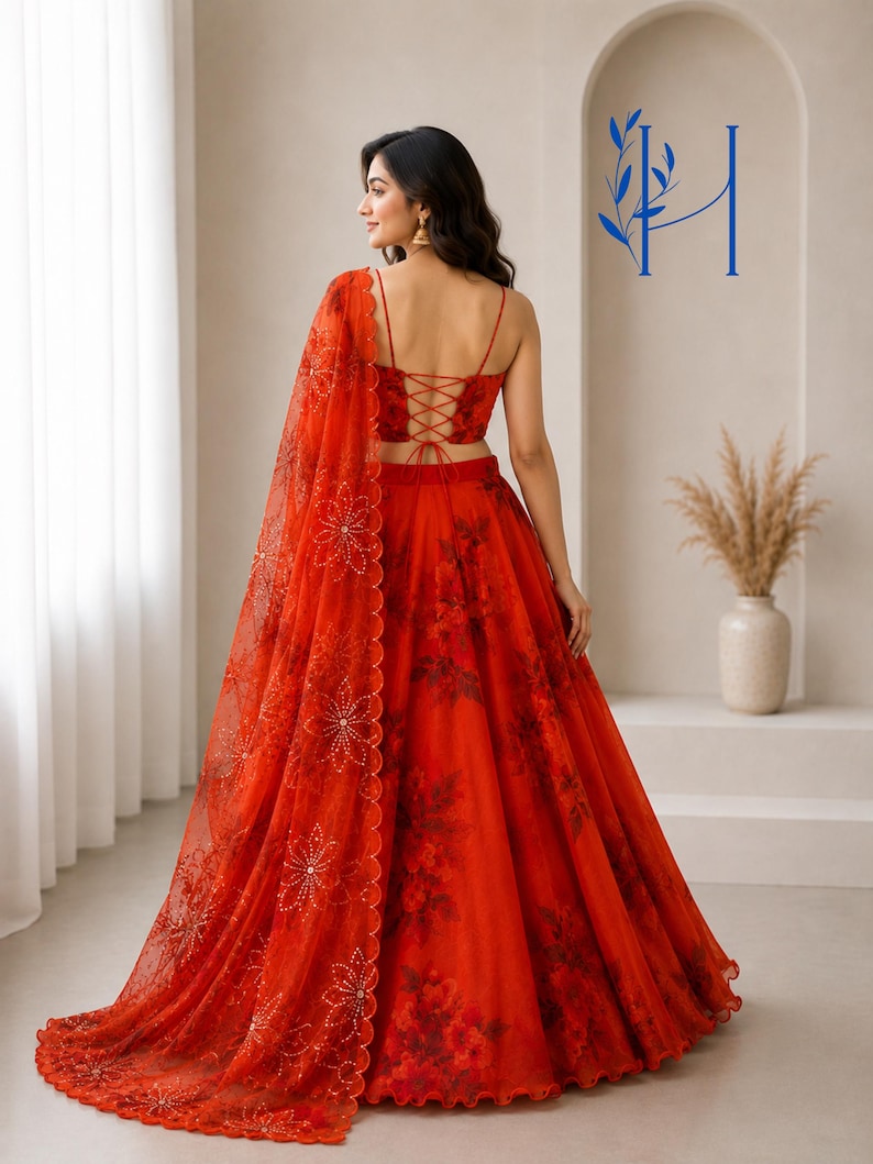 May include: A vibrant red lehenga set featuring a sleeveless crop top with a lace-up back and a full-length skirt with a dark floral print. A matching sheer red dupatta with embroidered floral patterns and scalloped edges drapes over the shoulder.