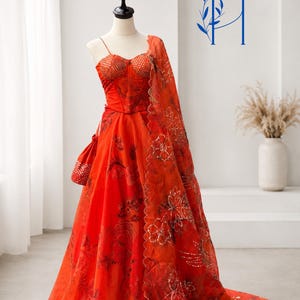 May include: A vibrant orange floor-length gown featuring a corset-style bodice with thin straps, a full skirt with dark floral patterns, and a matching sheer dupatta with scalloped edges and sequin embroidery, displayed on a mannequin.