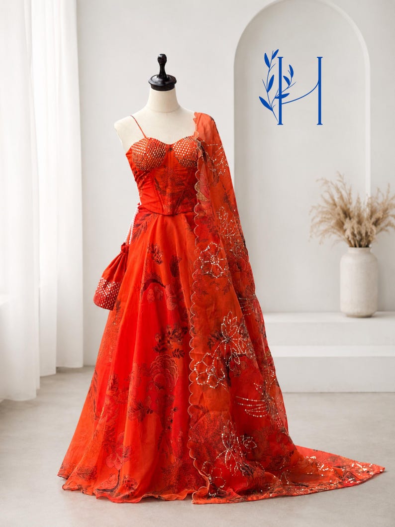 May include: A vibrant orange floor-length gown featuring a corset-style bodice with thin straps, a full skirt with dark floral patterns, and a matching sheer dupatta with scalloped edges and sequin embroidery, displayed on a mannequin.