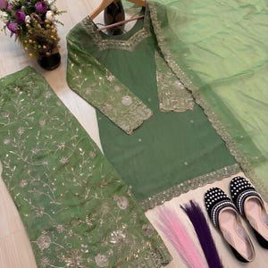 May include: A sage green embroidered outfit, including a tunic, pants, and a sheer scarf. The tunic has long sleeves and gold embellishments. Black and white beaded flats and colorful hair extensions are also present.