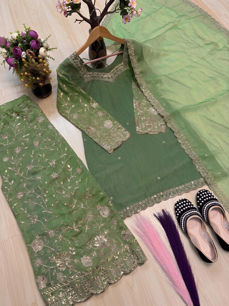 May include: A sage green embroidered outfit, including a tunic, pants, and a sheer scarf. The tunic has long sleeves and gold embellishments. Black and white beaded flats and colorful hair extensions are also present.