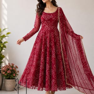 May include: A deep red floor-length dress with long sleeves and a round neckline, featuring a repeating white circular pattern throughout the fabric. The garment includes a matching sheer red dupatta draped over one shoulder with gold trim.