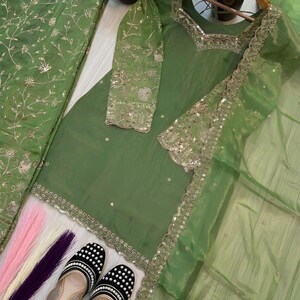 May include: A green embroidered salwar kameez set with matching dupatta. The set includes a tunic, pants, and a sheer scarf. The outfit is adorned with gold embroidery and sequins. Black and white beaded flats are at the bottom of the image.