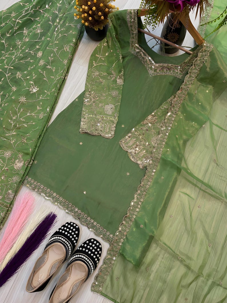 May include: A green embroidered salwar kameez set with matching dupatta. The set includes a tunic, pants, and a sheer scarf. The outfit is adorned with gold embroidery and sequins. Black and white beaded flats are at the bottom of the image.