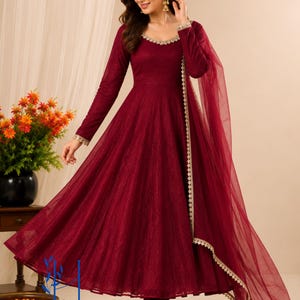 May include: A deep maroon floor-length Anarkali dress with long sleeves and a flared skirt. The neckline and sleeve cuffs feature intricate gold embroidery. A matching sheer maroon dupatta with a gold scalloped border drapes over the shoulder.