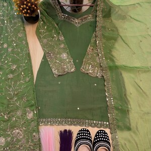 May include: An olive green salwar kameez set with intricate silver embroidery and embellishments. The set includes a long-sleeved tunic, matching pants, and a sheer dupatta. The image also shows a pair of black and white beaded shoes and decorative hair extensions.