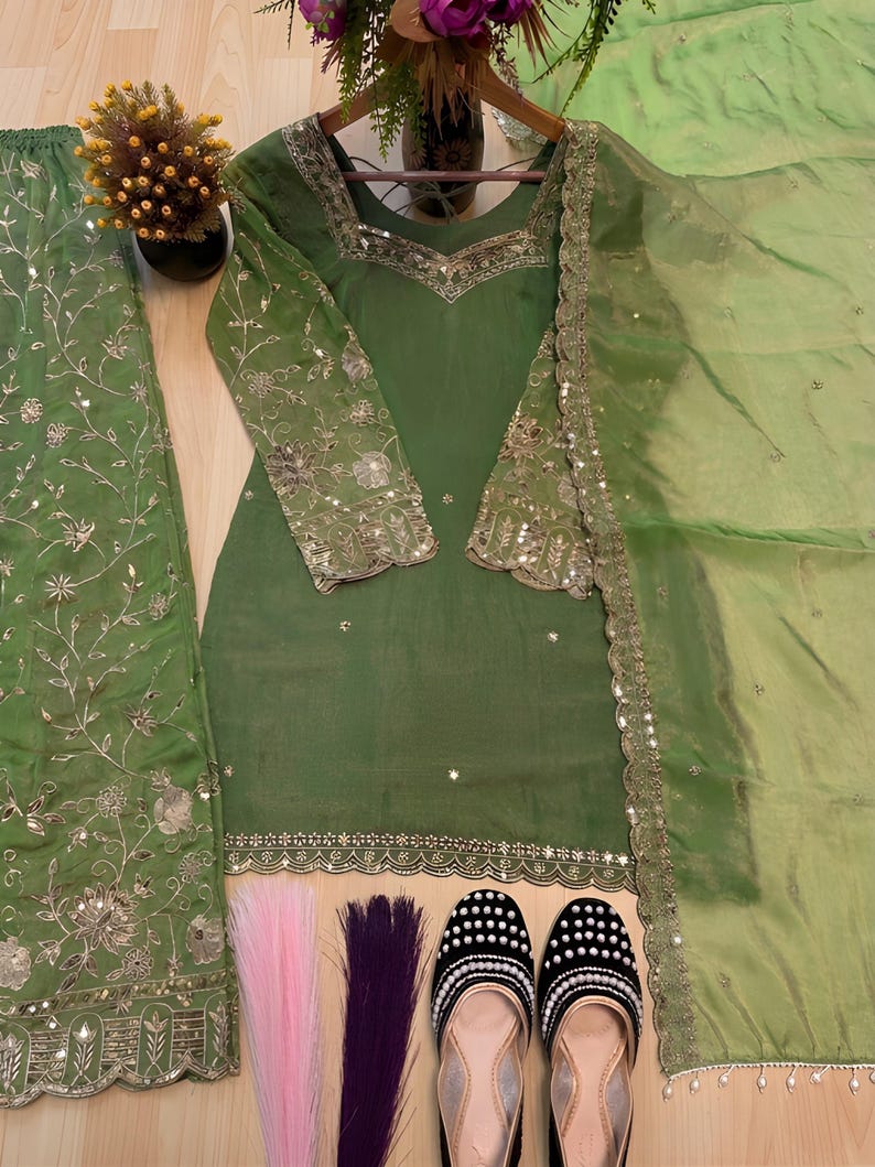 May include: An olive green salwar kameez set with intricate silver embroidery and embellishments. The set includes a long-sleeved tunic, matching pants, and a sheer dupatta. The image also shows a pair of black and white beaded shoes and decorative hair extensions.