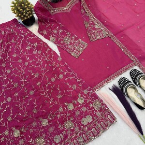 May include: A magenta-colored embroidered salwar kameez set. The set includes a long-sleeved tunic, wide-legged pants, and a matching scarf. Gold embroidery and mirror work adorn the garments. Black and white embellished shoes are also present.