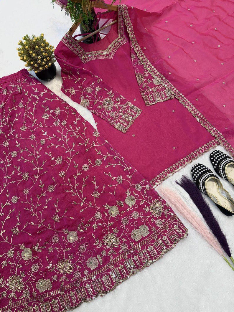 May include: A magenta-colored embroidered salwar kameez set. The set includes a long-sleeved tunic, wide-legged pants, and a matching scarf. Gold embroidery and mirror work adorn the garments. Black and white embellished shoes are also present.