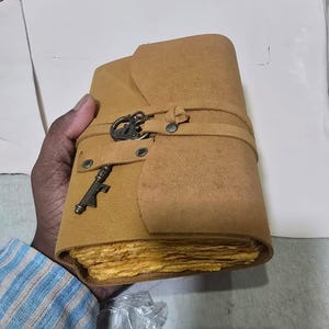 May include: A tan leather journal with a strap closure and a decorative key charm. The journal has a rustic, vintage aesthetic, with aged paper pages. The cover is a warm, earthy tone, suggesting a handcrafted item.