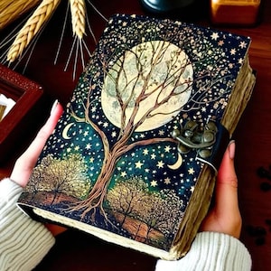 May include: A decorative journal with a tree of life design. The cover features a full moon, stars, and trees against a dark blue background. The journal is secured with a metal clasp and a leather strap.