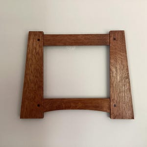 May include: A square wooden picture frame with a medium brown finish and visible wood grain. The frame features four straight sides with slightly flared vertical legs and small square dark wood inlays at each of the four corner joints.