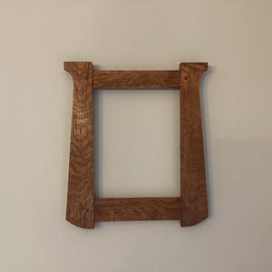 May include: A rectangular wooden picture frame with a medium brown finish and visible wood grain. The frame features two vertical side pieces that flare slightly at the top and bottom, connected by two horizontal crossbars to form an opening.