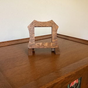May include: A small, square wooden display stand with a natural brown grain finish. The stand features a central open frame supported by two sturdy feet, designed to hold items upright. The wood exhibits a visible texture and warm brown tones.