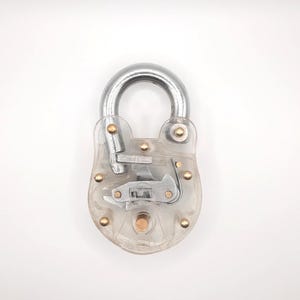 May include: A clear padlock with a silver shackle and internal mechanisms visible. The lock has a transparent body with brass-colored rivets. The design showcases the internal workings of a locking mechanism.