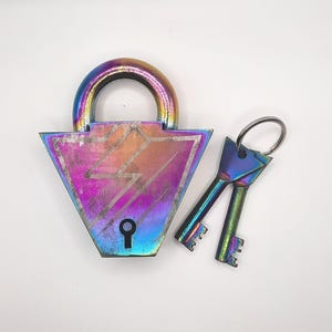 Puede incluir: Un candado decorativo y dos llaves con un acabado iridiscente de arcoíris. El candado tiene un cuerpo triangular con un diseño de rayo y un ojo de cerradura. Las llaves están unidas a un anillo de metal.