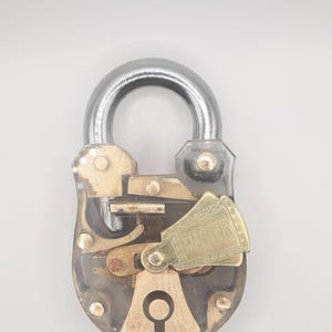 Puede incluir: Un candado antiguo con un grillete plateado y un cuerpo de latón. El mecanismo de la cerradura es visible, con intrincados componentes metálicos. La palabra "MASTER" está grabada en una placa de latón. El candado es para seguridad.