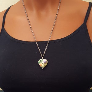 Könnte beinhalten: Eine silberne Kettenkette mit einem herzförmigen Anhänger. Der Anhänger ist weiß mit einem floralen Design in Grün-, Orange- und Lilatönen.