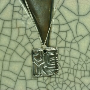May include: A silver necklace with a square pendant featuring an abstract design. The pendant has a textured surface and appears to be made of metal.